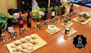 La imagen presenta una exhibición gastronómica con productos locales, probablemente de Los Realejos, en la isla de Tenerife. Sobre una mesa de madera, se observan botellas de vino, quesos, panes, conservas, además de otros alimentos como plátanos y hierbas frescas. En el centro de la mesa, las letras iluminadas "LOS REALEJOS" destacan el nombre del lugar o evento. Esta disposición sugiere una muestra de productos típicos de la región, lo que podría ser parte de una promoción de la gastronomía local. El logotipo en la parte inferior derecha refuerza el enfoque en la gastronomía de Los Realejos.