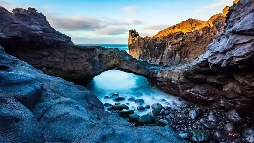 En la imagen se observa un espectacular paisaje costero en Tejina, con imponentes arcos naturales formados por la erosión en las rocas. Estos arcos se alzan majestuosamente sobre el océano, ofreciendo vistas panorámicas y un contraste dramático entre el verde de la vegetación y el azul profundo del mar. El cielo despejado y la luz del sol realzan la belleza natural del entorno, creando una escena impresionante y serena.