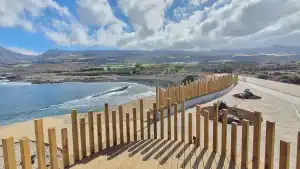 "Vista del paseo marítimo de Punta Blanca en Canarias, con un camino pavimentado bordeado de palmeras y edificios residenciales, y el océano azul al fondo bajo un cielo despejado."