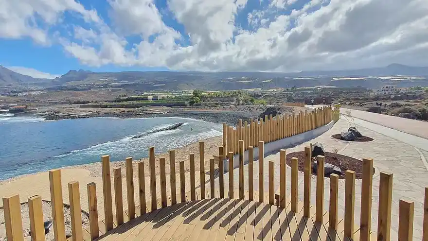 "Vista del paseo marítimo de Punta Blanca en Canarias, con un camino pavimentado bordeado de palmeras y edificios residenciales, y el océano azul al fondo bajo un cielo despejado."