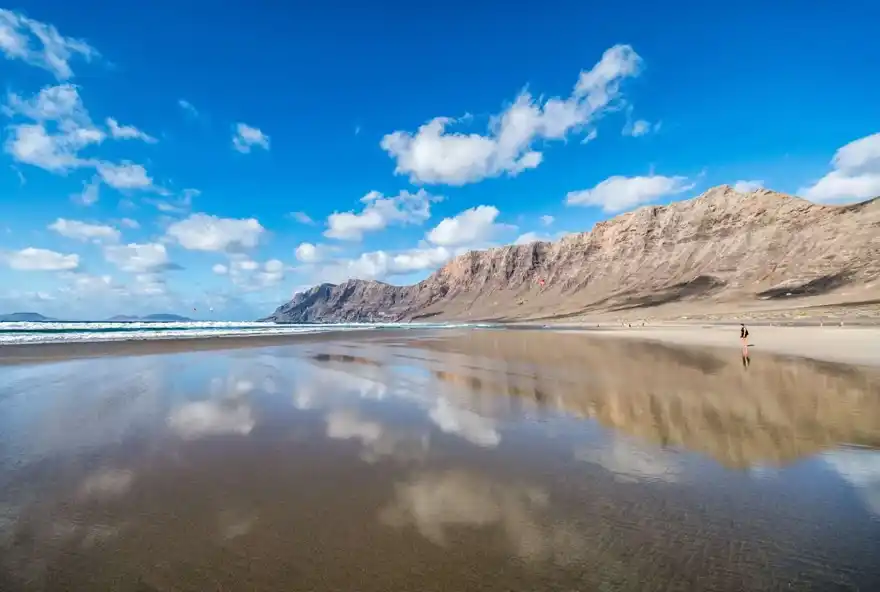 "Vista panorámica de la Playa de Famara, Lanzarote, con sus extensas arenas doradas y el impresionante macizo de Famara al fondo. Un destino ideal para los amantes del surf y la naturaleza en estado puro."