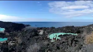 La imagen muestra la Fuente Santa en la isla de La Palma, Canarias. Es un balneario natural con piscinas termales rodeadas de rocas volcánicas. La ubicación está frente al océano Atlántico, lo que crea un paisaje impresionante donde las aguas termales contrastan con el entorno volcánico. Este lugar es famoso por sus propiedades curativas y su historia, ya que las aguas termales de la Fuente Santa fueron redescubiertas después de haber estado ocultas bajo lava durante siglos tras una erupción volcánica.
