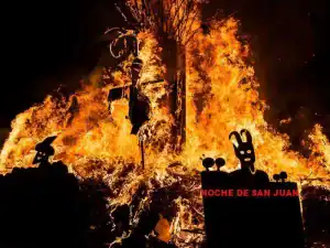 La imagen muestra una gran hoguera durante la celebración de la Noche de San Juan en la localidad de San Juan de la Rambla, Tenerife, con figuras y símbolos en primer plano mientras el fuego arde intensamente.