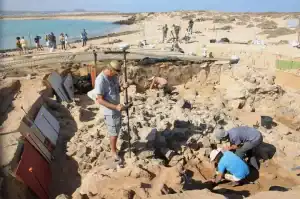 La imagen muestra un grupo de arqueólogos realizando excavaciones en un yacimiento en la costa de Fuerteventura, cerca del mar, donde se están descubriendo vestigios históricos relacionados con la presencia romana en la isla.