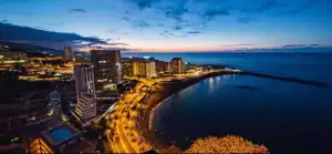 La imagen muestra una vista nocturna de una ciudad costera en Puerto de La Cruz, con edificios iluminados que se reflejan en el océano. El horizonte se tiñe con los últimos colores del atardecer, mientras las luces de la ciudad realzan el contraste entre la costa y el mar.