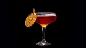 La imagen muestra una copa de vermut decorada con una rodaja de naranja deshidratada, sujeta al borde de la copa con un pequeño clip. La bebida tiene un tono ámbar rojizo y se presenta sobre un fondo oscuro, resaltando su elegancia y sofisticación.