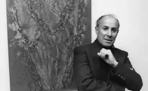 Fotografía en blanco y negro de César Manrique, reconocido artista y arquitecto canario, posando junto a una de sus obras abstractas.