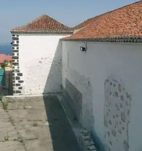 La imagen muestra una parte exterior del antiguo castillo de San José, una estructura histórica en Canarias, con paredes blancas y detalles de piedra negra en las esquinas, junto a un techo de tejas rojas.