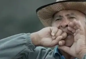 La imagen muestra a un hombre mayor utilizando el "silbo gomero", un lenguaje silbado tradicional de la isla de La Gomera, en Canarias. Lleva un sombrero de paja y parece estar en un entorno rural mientras ejecuta este medio de comunicación ancestral.