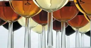 La imagen muestra varias copas de vino dispuestas en fila. Las copas contienen vinos de diferentes colores, que varían desde tonalidades doradas claras hasta tonalidades más oscuras como ámbar y rojo intenso. Estas diferencias de color reflejan la variedad de tipos de vino, posiblemente desde blancos y dorados hasta tintos y vinos dulces, típicos de la rica tradición vitivinícola de las Islas Canarias. Las copas destacan por su elegancia y la luz que se refleja a través del vino.