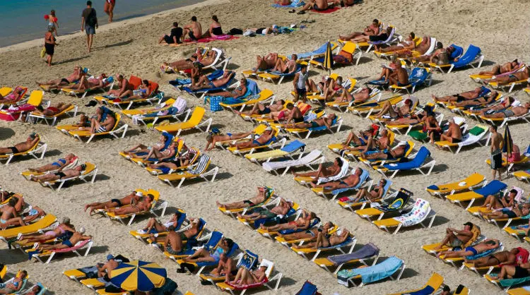 "Turistas disfrutando del sol en tumbonas, alineados en la playa en la costa de las Islas Canarias."