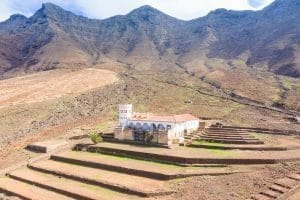 Edificio colonial con torre, arcos y techo rojo, rodeado por montañas escarpadas y una cerca de piedra.