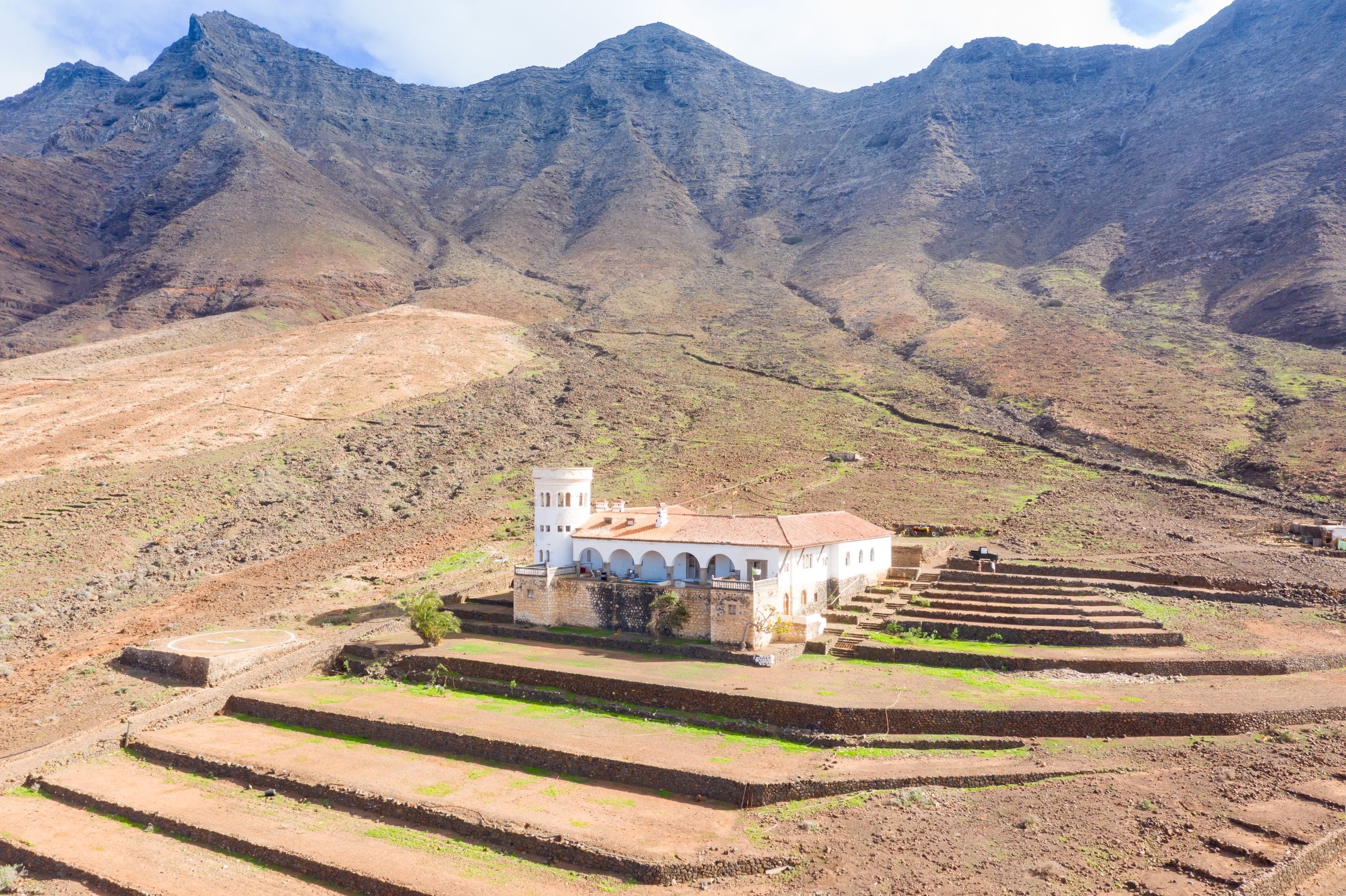 Edificio colonial con torre, arcos y techo rojo, rodeado por montañas escarpadas y una cerca de piedra.