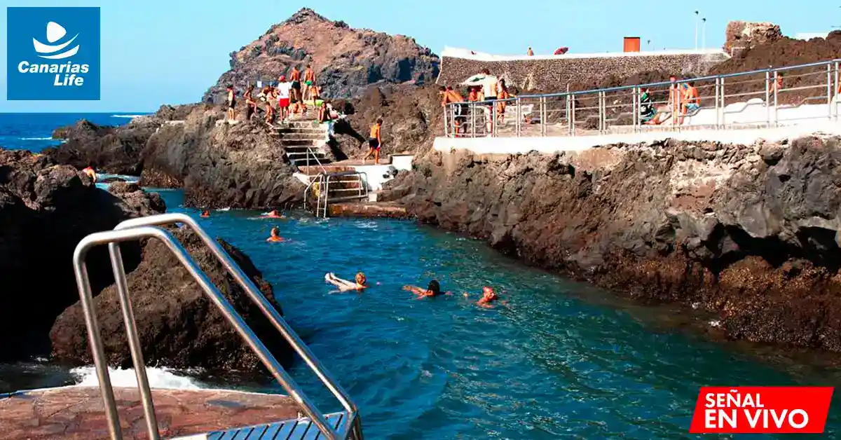 "Canarias Life" muestra una playa con gente nadando en aguas cristalinas, rodeada de rocas y montañas, con un ambiente relajado y turístico.