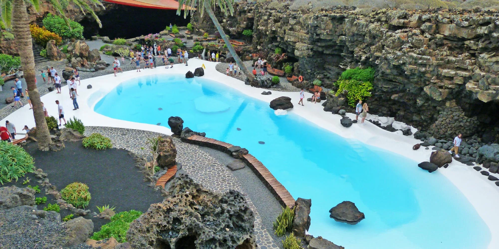 "Vista interior de los Jameos del Agua en Lanzarote, mostrando un espacio subterráneo con una piscina natural de agua cristalina, rodeada de formaciones rocosas volcánicas y vegetación exótica bajo un techo parcialmente abierto que permite la entrada de luz natural."