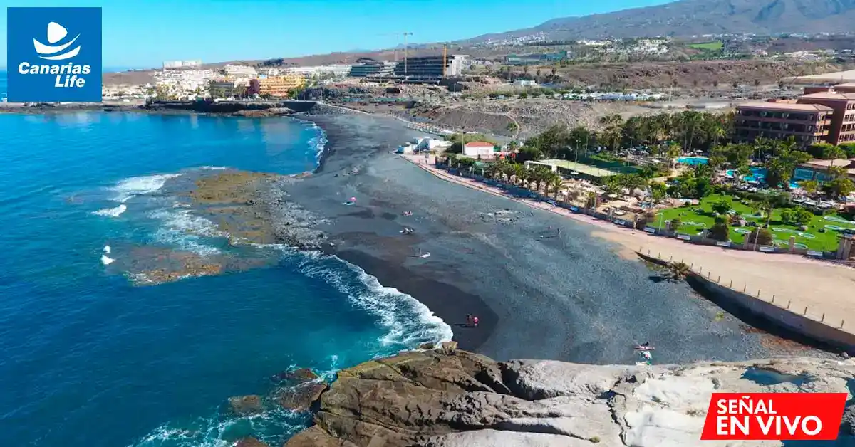 Playa con arena negra, edificios modernos, montañas en el fondo, mar azul, y gente disfrutando del paisaje.