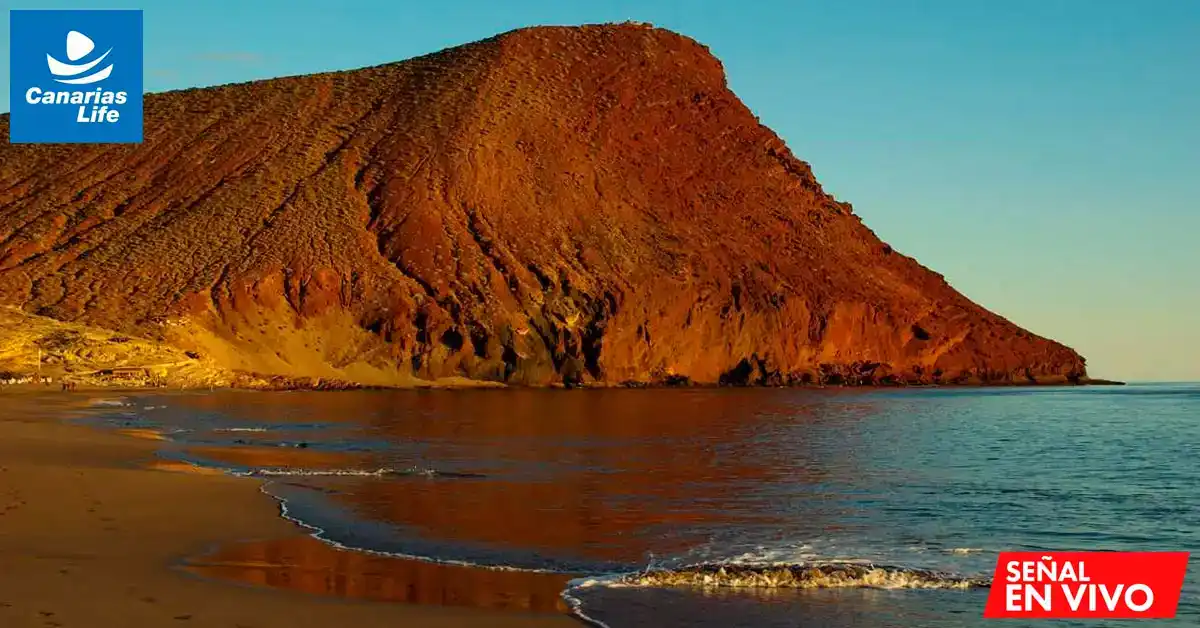 "Playa con montaña rocosa, mar azul, y sol en la costa de Tenerife."