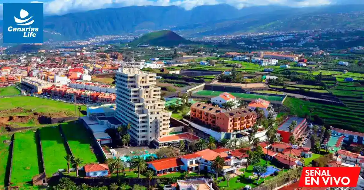 Panorámica de una ciudad costera con edificios modernos, campos verdes y montañas en el fondo, bajo un cielo azul.