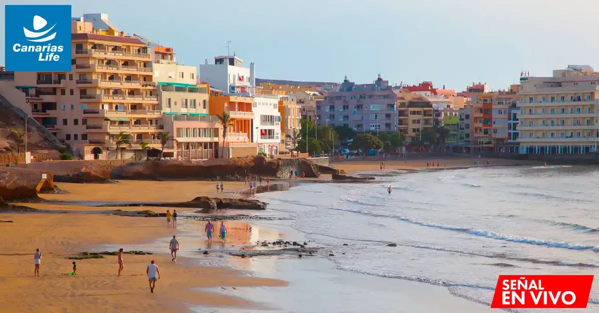 Playa con edificios coloridos, gente caminando y nadando, mar y sol, Canarias Life logo.