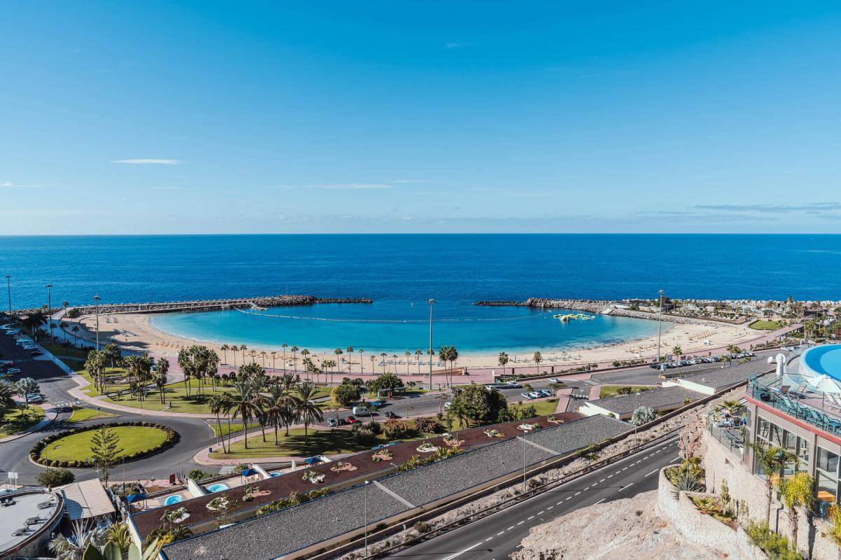 Vista panorámica de una playa turística en Gran Canaria, con aguas tranquilas y claras, arena dorada y numerosas personas disfrutando del sol, rodeada por montañas.