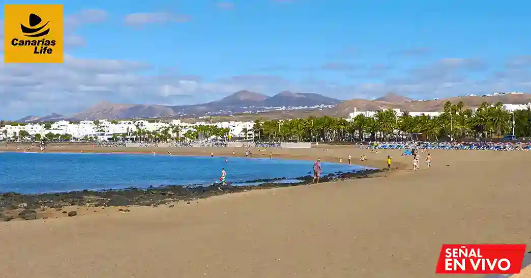 Platja con arena blanca, agua azul, montañas en el fondo, playas llenas de gente, palmeras y edificios blancos.