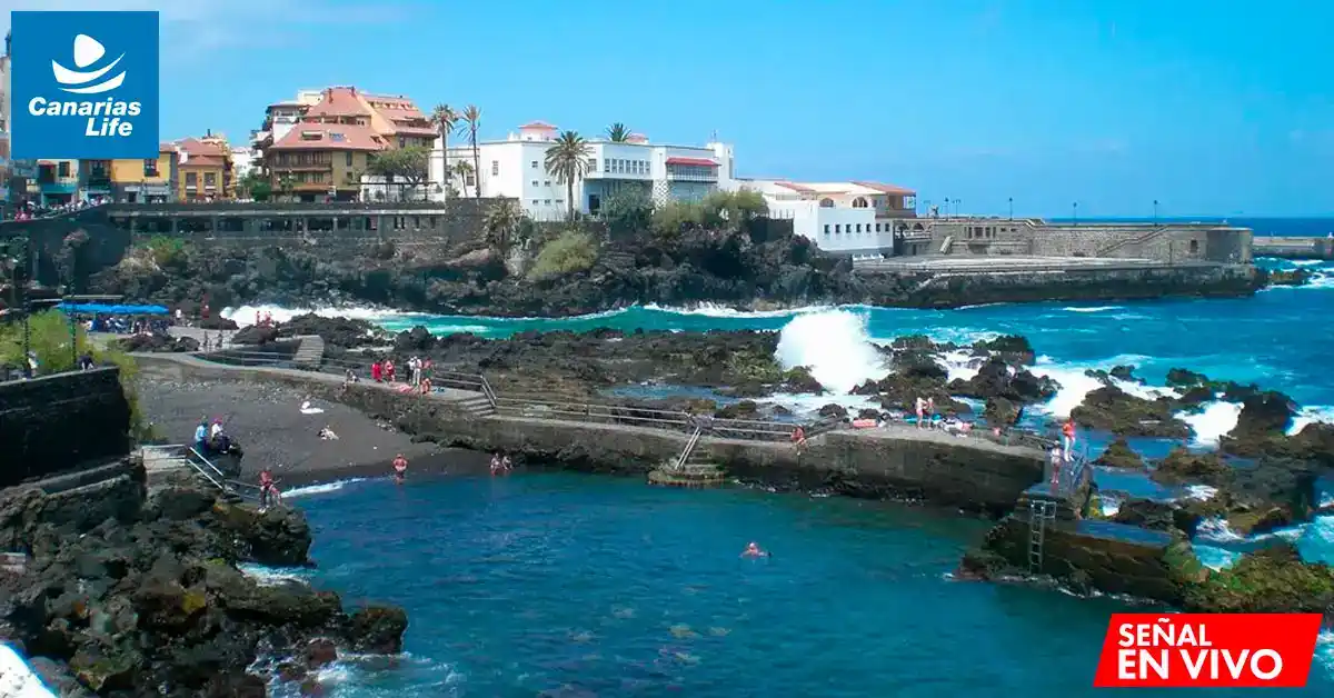 Playa con olas, edificios costeros, y gente disfrutando del agua bajo un cielo azul.