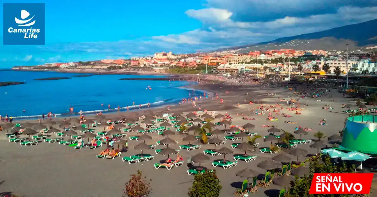 Plage animée avec chaises et parasols, vista desde arriba, playas de arena negra, mar azul, montaña en el fondo, cielos azules con nubes.
