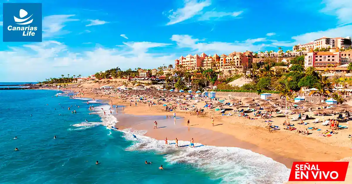 Playa concurrida con edificios, arena dorada, agua azul y sol brillante; turistas disfrutando del clima cálido.