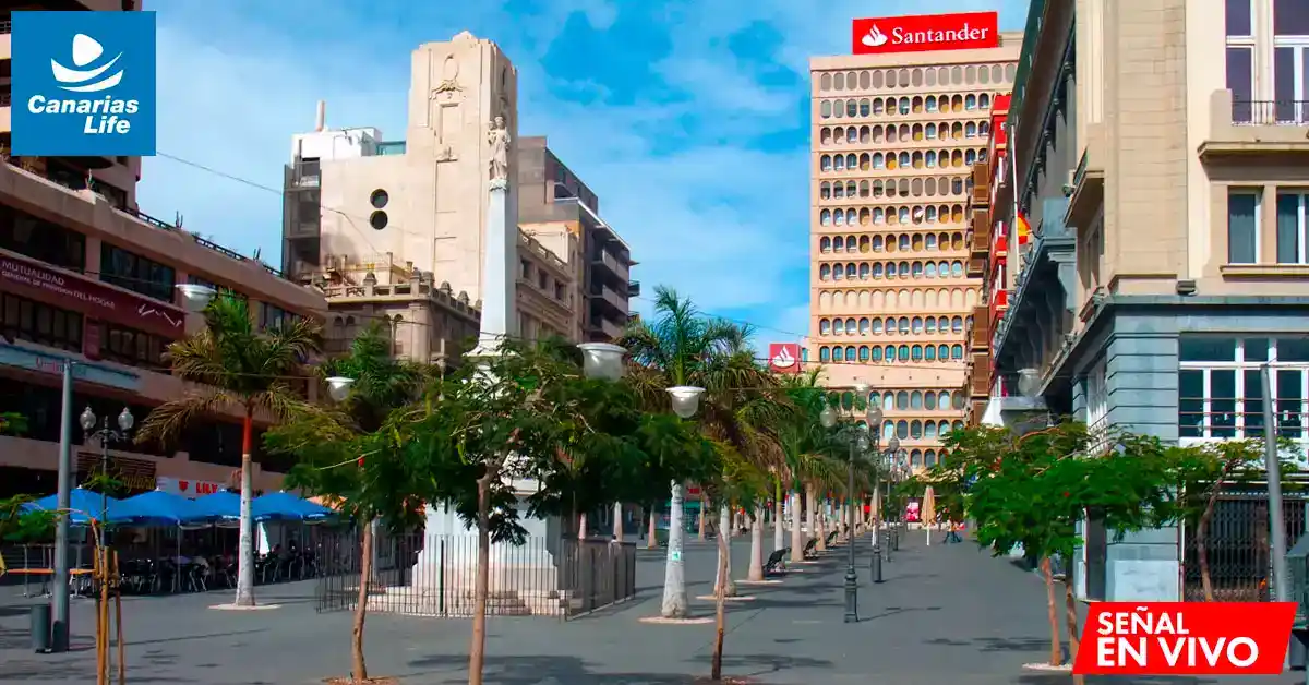 Plaza con monumento central, edificios modernos y comerciales, árboles y sombrillas en la terraza de un restaurante.