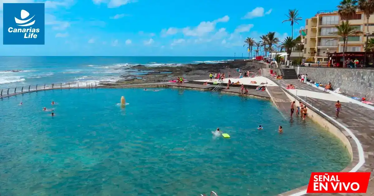 Piscina natural en la costa, con gente disfrutando del agua y el sol bajo un cielo azul despejado.