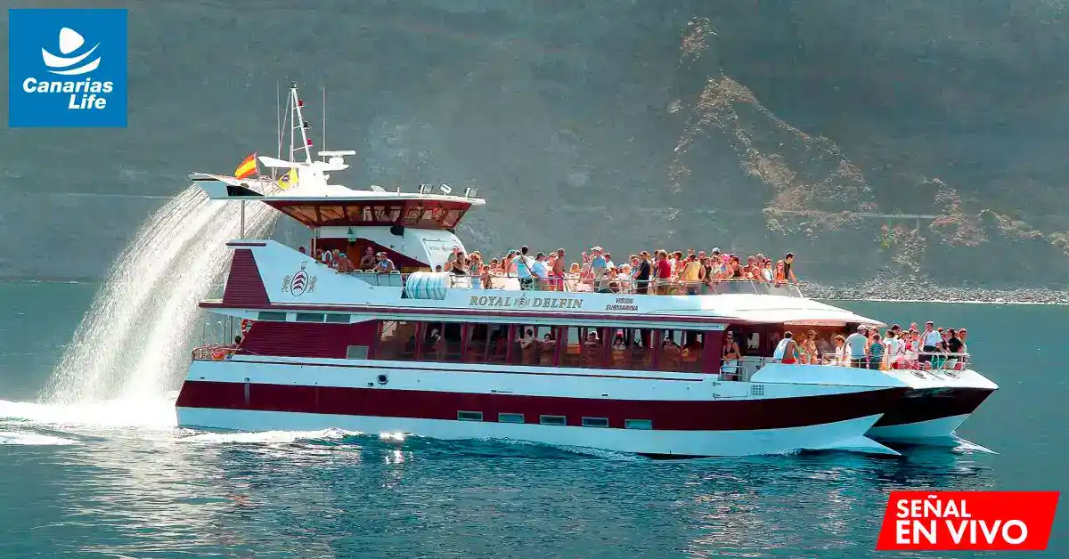 Barco "Royal Delfín" con pasajeros en el agua, bandera española, paisaje montañoso, logo Canarias Life, señal en vivo.
