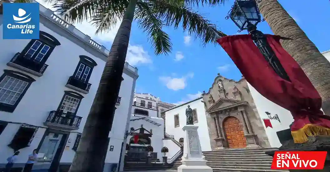 Plaza colonial con edificios blancos, palmeras, estatuilla central, y una iglesia histórica en el fondo.