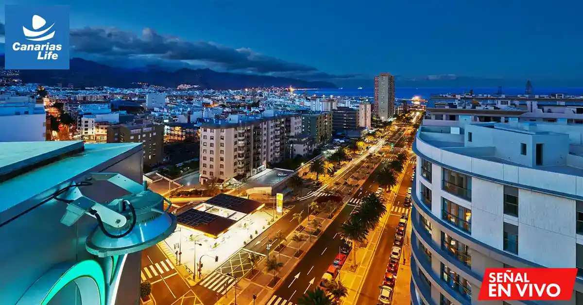Panorámica nocturna de una ciudad costera con edificios, calles iluminadas y mar visible en el horizonte.