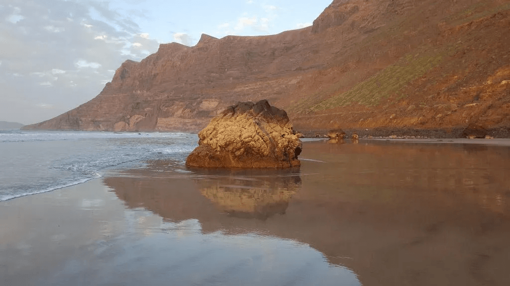 Famara Lanzarote. Playa con roca central, reflejo en la arena, montañas en el fondo, mar tranquilo, atardecer.