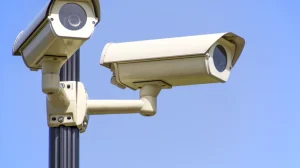 Una imagen muestra un primer plano de dos cámaras de tráfico instaladas en un poste metálico. Las cámaras, de color gris oscuro, están orientadas hacia la carretera. Una está ligeramente más arriba que la otra. Detrás de las cámaras se aprecia un cielo nublado y parcialmente azul. La imagen parece ser representativa del monitoreo del tráfico en las carreteras.