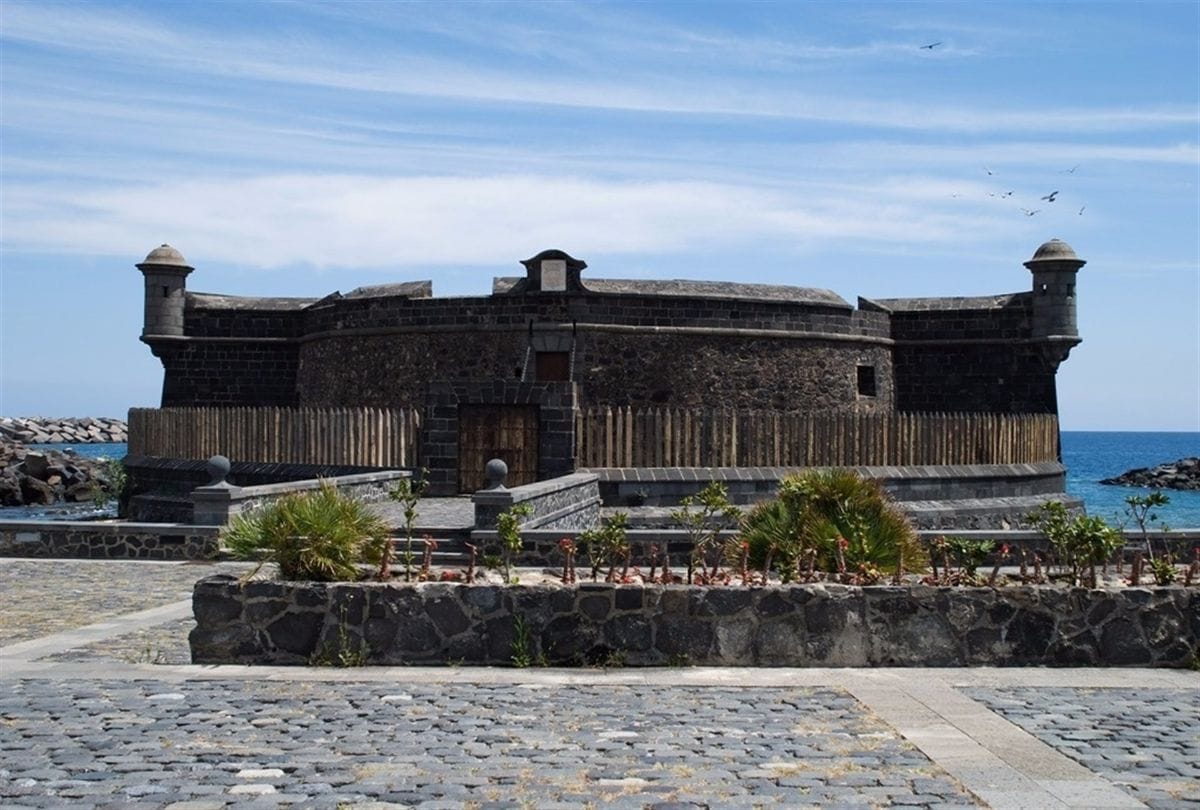 Castillo de San Cristóbal Tenerife - Fortaleza histórica con torres, construida en piedra negra, rodeada por una cerca de madera y ubicada cerca del mar, bajo un cielo azul despejado.