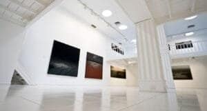 Galleria d'arte moderna con dipinti astratti, colonne bianche, illuminazione circolare, e vetrate al secondo piano.