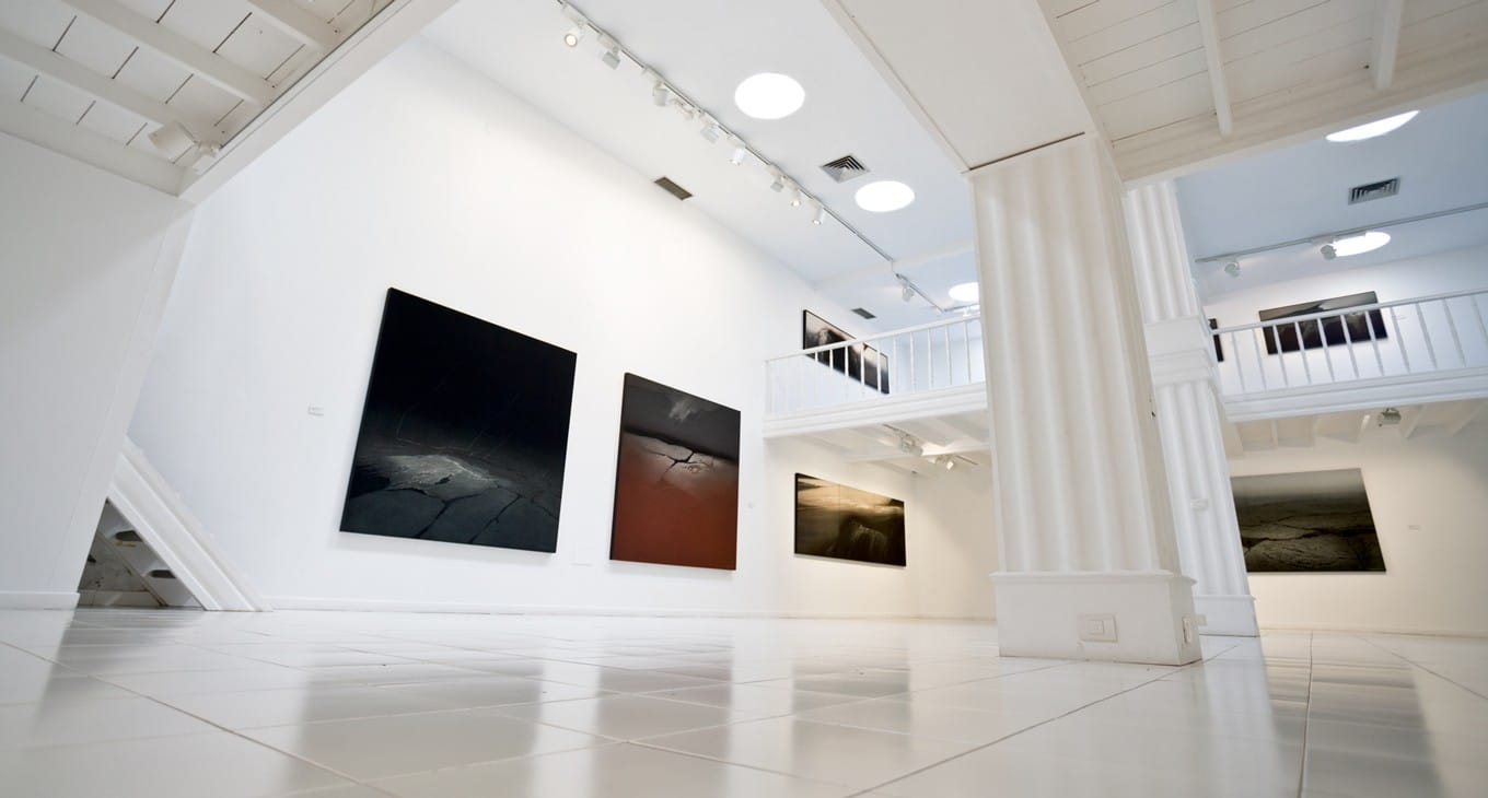 Galería de arte moderna con pinturas abstractas, columnas blancas, iluminación circular, y acristalado en la segunda planta.