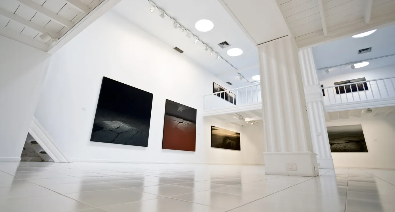Galería de arte moderna con pinturas abstractas, columnas blancas, iluminación circular, y acristalado en la segunda planta.