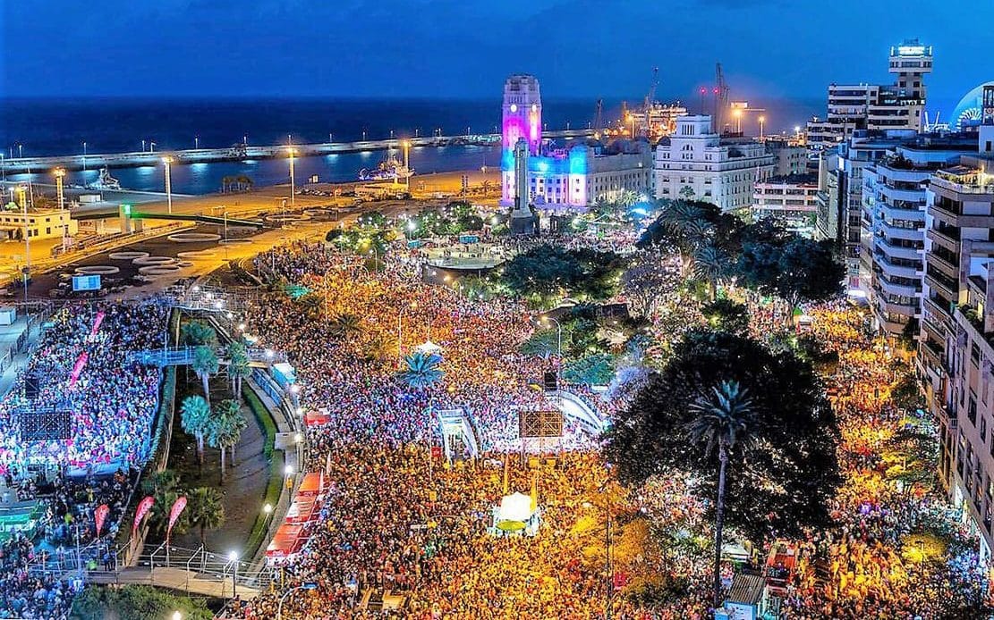 Carnaval de Tenerife, Festival nocturno vibrante con gran multitud, iluminación colorida, y paisaje urbano costero.