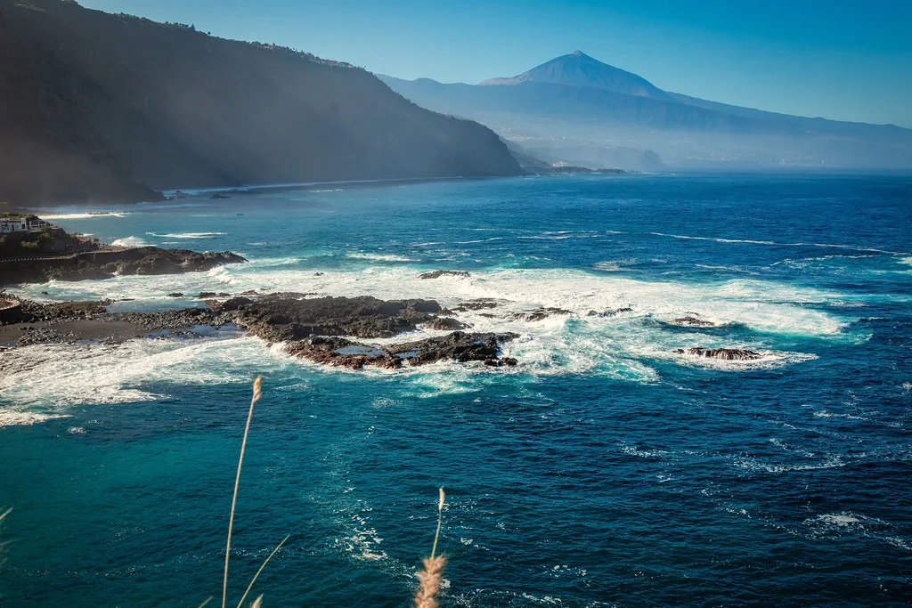 "Costa rocosa con olas violentas, montaña distante bajo un cielo azul claro."