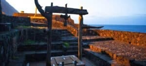 Antico cortile con gradini, muro di pietra e arco di legno, con vista sul mare al tramonto.