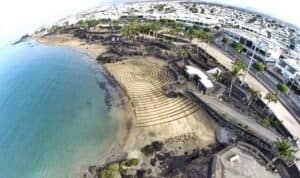 "Disegno a spirale sulla sabbia vicino alla spiaggia, con edifici e palme sullo sfondo."
