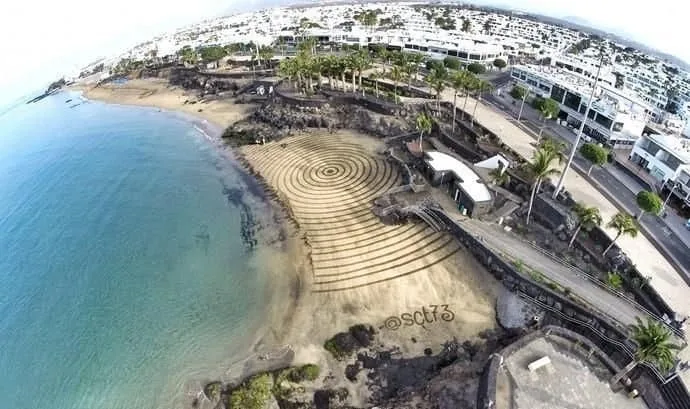 "Spiral design in the sand near the beach, with buildings and palm trees in the background."