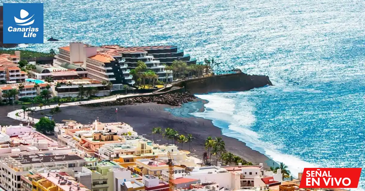 "Canarias Life" muestra una impresionante vista aérea de una playa con edificios modernos, contrastando con la naturaleza salvaje del mar azul brillante y las olas rompiendo en la orilla negra arenosa.