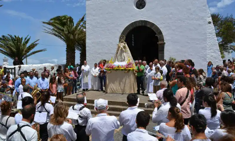 Fiesta religiosa con statue de la Virgen, música y congregación en una iglesia blanca.