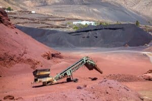 Miniera industriale con macchinari pesanti, cumuli di rocce nere e rosse, e edifici in lontananza.