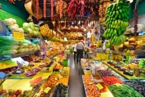 Mercato pieno di frutta e verdura fresche, con banane appese, cipolle bianche, pomodori rossi e altri prodotti agricoli.