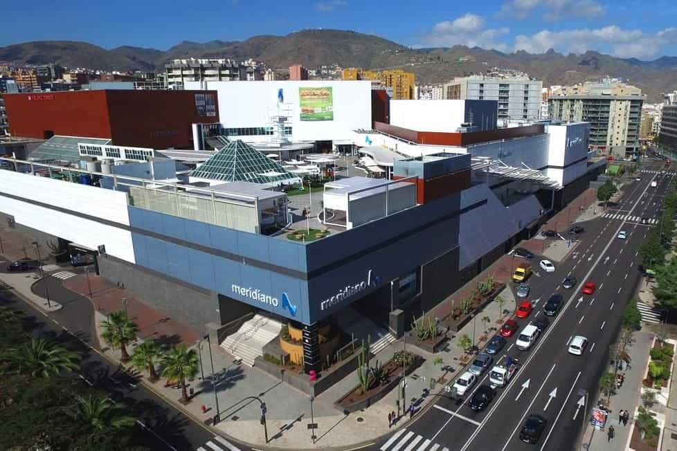 Centro comercial modernista con tiendas, parking y accesos directos a la calle, rodeado de edificios residenciales y montañas en el horizonte.