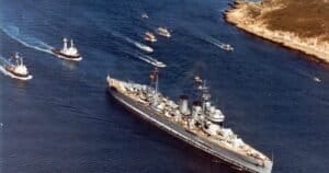 Un' storica nave da crociera naviga sull'acqua, scortata da vari barche più piccole, con una costa rocciosa sullo sfondo.